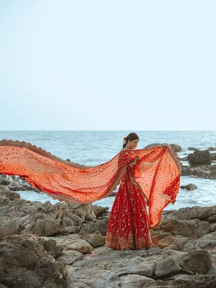 The bride lets her vibrant red dupatta fly in the wind, creating a dynamic and breathtaking shot against the ocean. This image is full of life and movement.