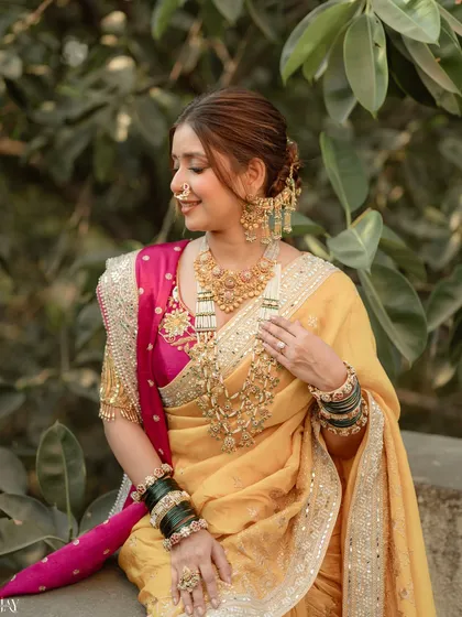 A beautiful seated portrait of the bride, showcasing her elaborate gold necklaces, green bangles, and the rich texture of her yellow and pink saree. Her serene expression adds to the elegance of the photo.