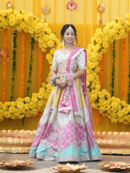 A full-length shot of the bride against a marigold backdrop. Her makeup is soft and elegant, complementing her beautiful multi-colored lehenga.