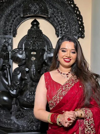 A happy bride posing in front of a Ganesha idol, her red saree and radiant makeup looking perfect for a post-wedding ceremony.