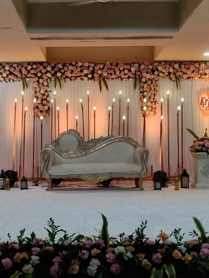 I designed this elegant engagement stage for an indoor hall. The setup features a classic white chaise lounge, a backdrop of warm candle-style lights, and a full floral border, creating a romantic and sophisticated look.