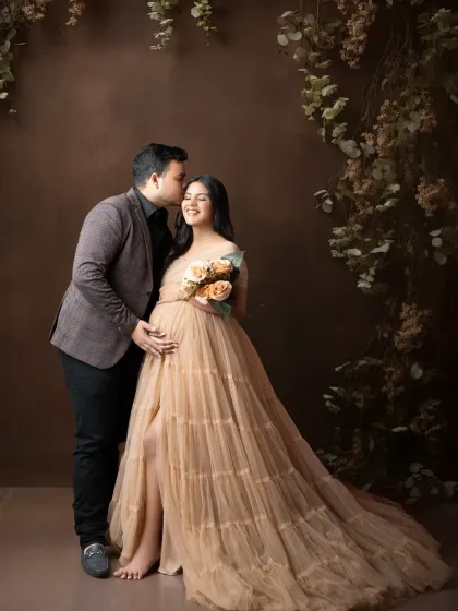An intimate and moody maternity portrait. The rich, dark brown backdrop and delicate floral accents create a warm, romantic feel, highlighting the tender connection between the expecting parents.