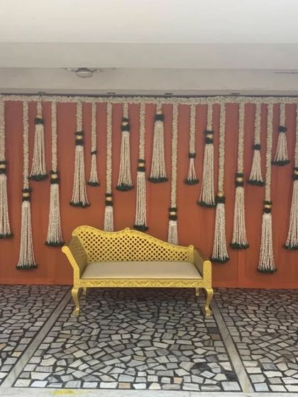 A simple yet elegant photo spot featuring a backdrop of hanging jasmine tassels against a vibrant orange wall. The golden chaise lounge provides a perfect place for guests to take memorable pictures.