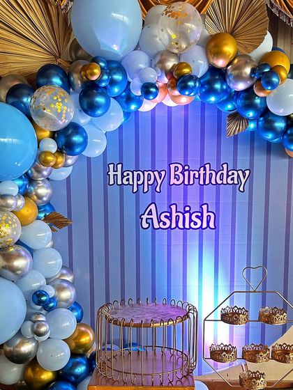 A handsome blue, silver, and gold themed birthday setup for Ashish. The decor features a striped backdrop and an elegant balloon arch with confetti and chrome balloons.