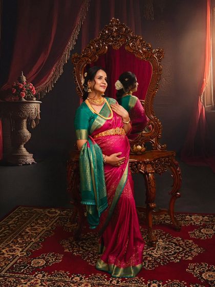 Reflecting on the journey ahead. This portrait, set in a room with regal, vintage decor, captures a moment of quiet contemplation, with the rich pink and green Kanjivaram saree adding a touch of royal elegance.