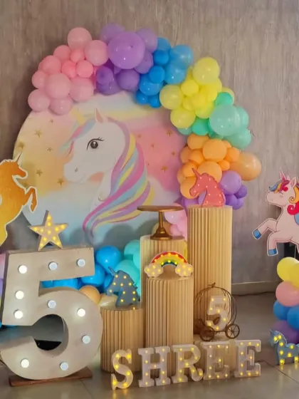 A magical unicorn-themed 5th birthday party. The centerpiece is a large unicorn backdrop surrounded by a pastel rainbow balloon garland, with unicorn cutouts and a light-up number '5'.