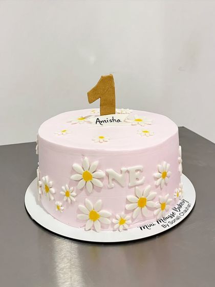 A pretty and delicate cake for a one-year-old girl. The design features a soft pink frosting covered in hand-painted daisies and a simple number '1' topper.