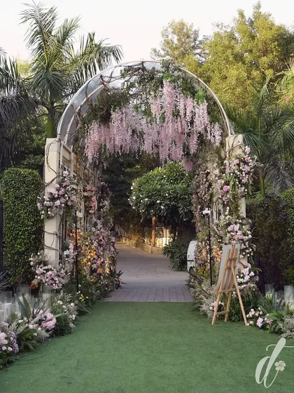 A grand floral archway serves as a magical entrance for a romantic wedding. Draped in cascading pink wisteria and lush greenery, this entryway immediately sets a tone of enchantment and refined elegance for arriving guests.
