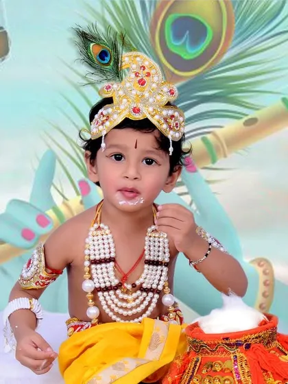 A toddler dressed as Krishna, complete with a peacock feather, flute, and a pot of butter. The custom illustrated backdrop adds a magical touch to these festive portraits.