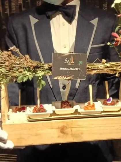 Our server presents a tray of Bhuna Ananas, a smoky and sweet grilled pineapple appetizer. The rustic wooden tray adorned with fresh flowers and herbs demonstrates our unique and thoughtful presentation style.