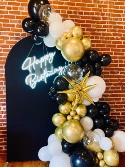 A modern and chic birthday setup with a black arched backdrop. The decoration is accented with a black, gold, and white balloon garland and a "Happy Birthday" neon sign.