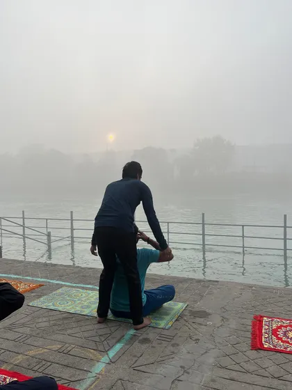 Assisting a student with a seated stretch, with the morning sun rising over the misty river in the background.