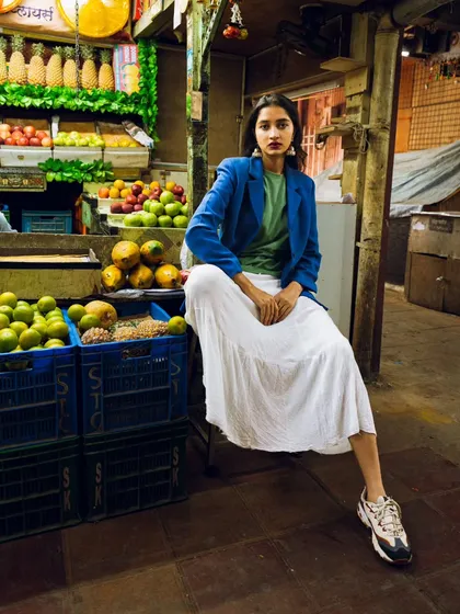A full-length shot showcasing the entire outfit, from the modern blazer to the sneakers, against the rustic backdrop of a market stall.