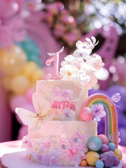 A beautiful two-tiered cake for Ayra's first birthday, decorated with delicate butterflies, a rainbow, and pastel-colored spheres.