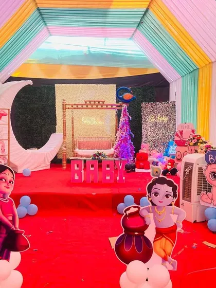 A baby shower stage inside a colorful pandal, decorated with character cutouts, a moon prop, and a traditional swing.