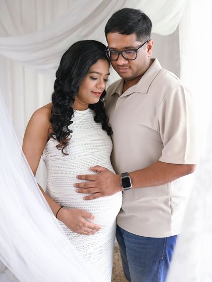 A tender moment between the expecting couple in a bright, all white studio setting. The soft light and gentle embrace create a feeling of peace and love.