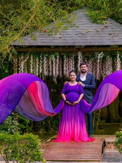 A powerful maternity shot on the wooden bridge, with the flowing purple gown creating a wing-like effect against the garden backdrop.