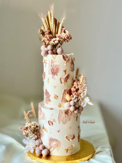A two-tiered boho-themed birthday cake in a palette of brown, beige, and off-white. It's decorated with dried florals, edible chocolate spheres, and textured buttercream for a rustic yet chic look.