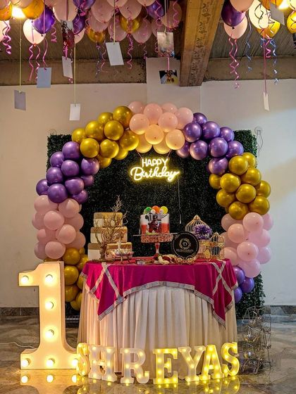 A first birthday celebration for Shreyas, featuring a large light-up '1' and his name in marquee letters. The scene is framed by a purple, pink, and gold balloon arch against a green hedge backdrop, creating a vibrant and personal setting.