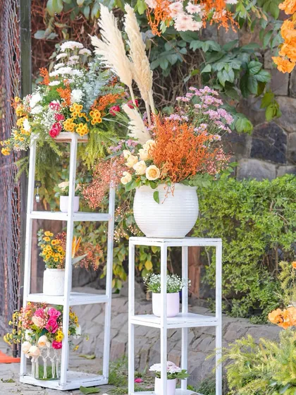 A beautiful floral corner with arrangements on white tiered shelves, featuring a mix of pampas grass, peach roses, and burnt orange foliage.
