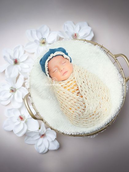 This baby boy, in a cream-colored swaddle and blue bonnet, looks like a precious pearl surrounded by white flowers in a golden basket.