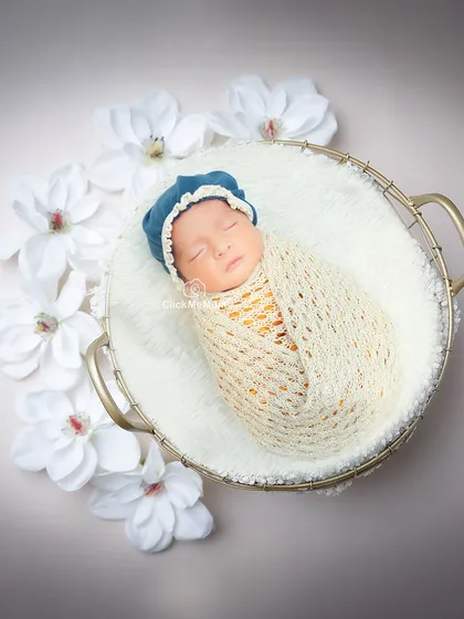 This baby boy, in a cream-colored swaddle and blue bonnet, looks like a precious pearl surrounded by white flowers in a golden basket.