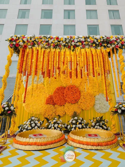 A classic Haldi ceremony setup, featuring a backdrop of marigold garlands, traditional low seating (urlis), and a bright yellow and white chevron floor.