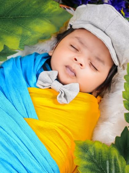 A newborn wrapped in vibrant blue and yellow, resting among lush green leaves. The flat cap and bow tie add a playful and stylish touch to this colorful setup.