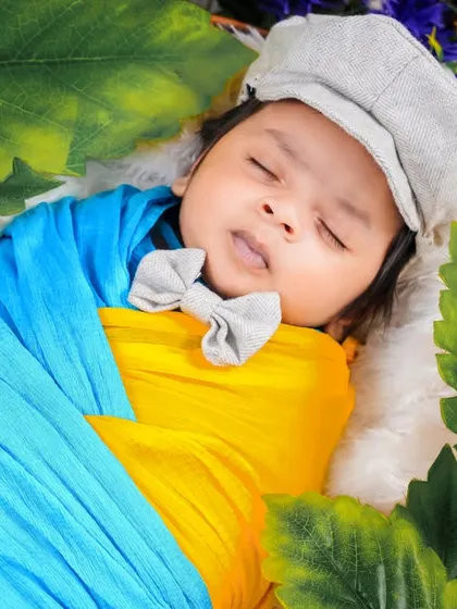 A pop of color with this baby wrapped in bright blue and yellow, wearing a little grey cap and bow tie, nestled amongst green leaves.