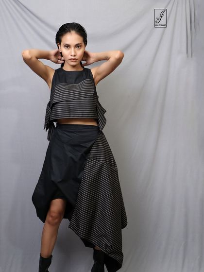 A model showcases an avant-garde, deconstructed outfit against a simple grey studio backdrop. The pose is strong and fashion-forward.