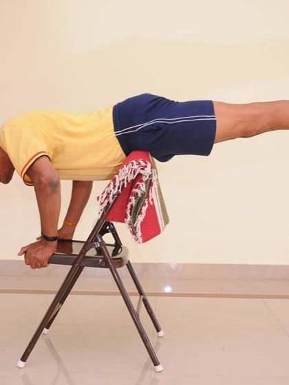 Mayurasana (Peacock Pose) supported by a chair. The chair bears some of the body's weight, allowing you to build the immense core and arm strength required for this advanced arm balance.