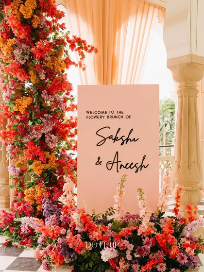 A beautiful welcome sign for a "Flowery Brunch" in Jaipur. The soft pink sign is framed by a stunning, overflowing arch of bougainvillea and other flowers in shades of pink, orange, and red.