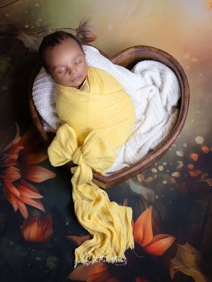 A beautiful autumn-themed shot with a baby in a yellow wrap inside a heart-shaped bowl, set against a backdrop of sunflowers.