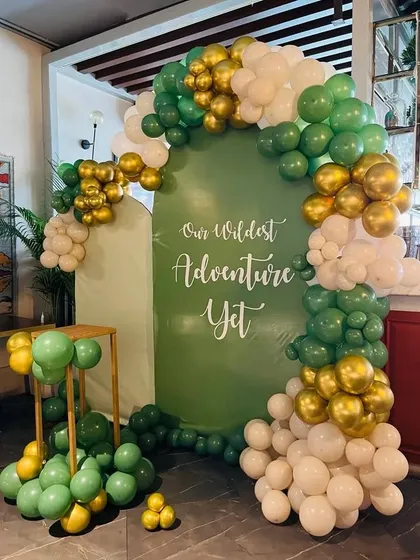 A gorgeous backdrop for a jungle or adventure-themed baby shower. The custom sign reads 'Our Wildest Adventure Yet', framed by an organic balloon arch in shades of green, white, and gold.