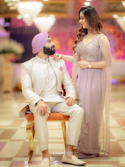 A regal and stylish portrait of the couple. Sanjit is seated while Simran stands beside him, her hand gently on his shoulder, creating a look of classic elegance and connection.