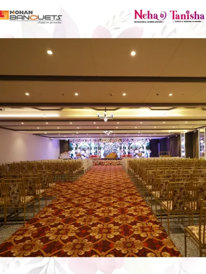 A view down the main aisle of Mohan Banquets, showcasing the expansive, patterned carpet and rows of seating leading towards a beautifully decorated stage, ready for a grand ceremony.