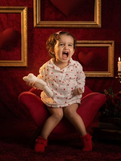 A toddler makes a fierce expression while sitting on a red chair in a dramatic, red velvet-themed studio setup with candles.