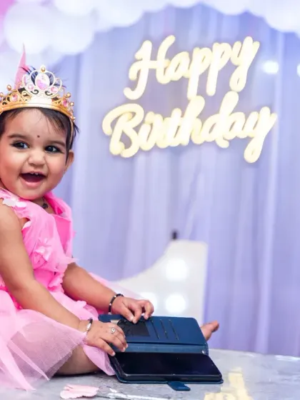 The birthday girl, dressed as a little fairy, sits happily at a table in front of a 'Happy Birthday' sign and balloon arch.