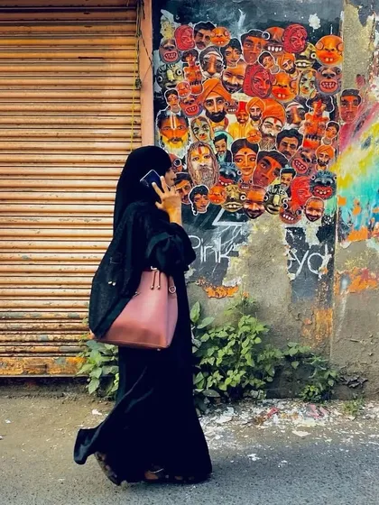 A woman in a hijab walks past the collaborative paste-up wall, talking on her phone. The image captures the intersection of daily life and street art in a busy Mumbai lane.