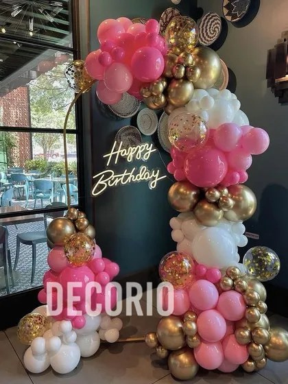 A stylish balloon arrangement in a restaurant setting, featuring a half-arch of pink, white, and gold balloons. The 'Happy Birthday' neon sign adds a celebratory glow to the space.