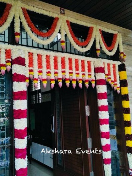 A traditional floral decoration for a housewarming ceremony. The main door is beautifully framed with garlands of red, white, and yellow flowers, creating a warm and festive welcome.