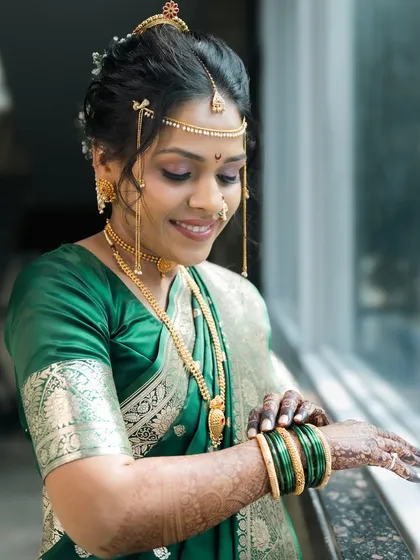 The beautiful bride Shraddha, dressed in a green Paithani saree, adjusts her bangles while looking out a window.