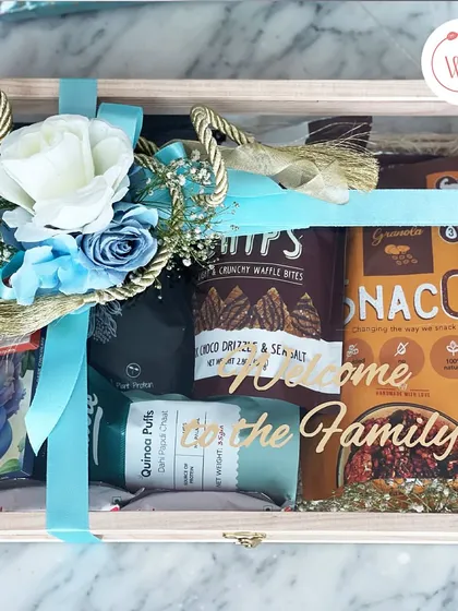A "Welcome to the Family" snack hamper in a wooden box with a clear lid, filled with healthy treats like quinoa puffs and granola.
