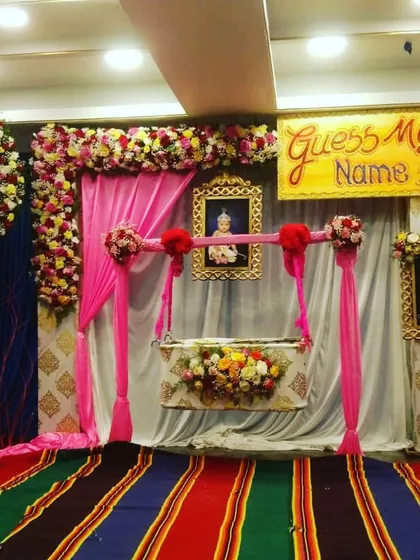 A grand and traditional naming ceremony setup. The decor includes a decorated cradle, pictures of baby Krishna, and a "Guess My Name" sign, all set against rich pink and blue drapes.