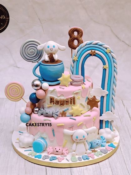 A dreamy two-tier Cinnamoroll-themed cake. This chocolate cake is decorated in a sweet pastel pink and blue palette, featuring the cute character, a fondant rainbow, and candy-like details.