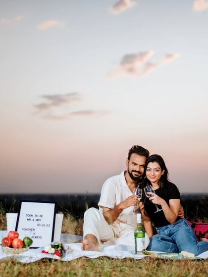 A romantic picnic setup at sunset for a pre-wedding shoot. The couple shares a toast, creating a beautiful and intimate scene against the evening sky.