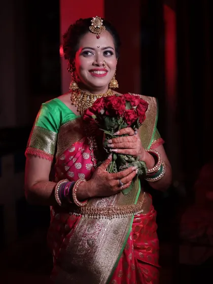 A happy bride holding a bouquet of roses. The red of the roses picks up the tones in her saree and makeup.