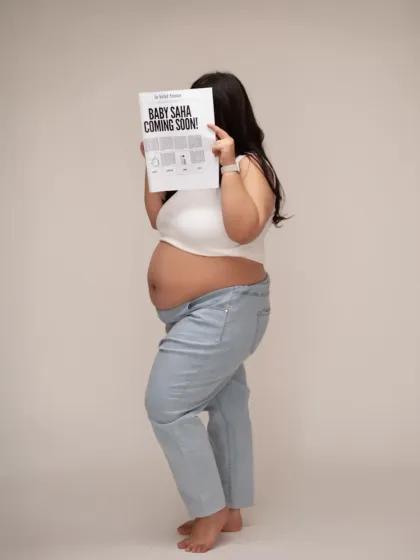 A creative and modern baby announcement. The mother-to-be holds a custom newspaper headline reading "Baby Saha Coming Soon!", a unique and fun way to share the news.
