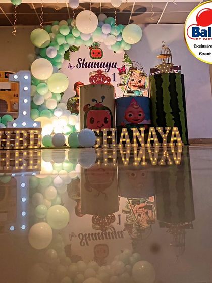A full-stage Cocomelon-themed first birthday. The decor includes a pastel balloon arch, the iconic yellow bus, and a 'SHANAYA' marquee.