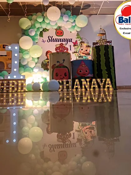 A full-stage Cocomelon-themed first birthday. The decor includes a pastel balloon arch, the iconic yellow bus, and a 'SHANAYA' marquee.